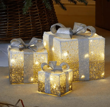 Three silver glitter gift boxes with matching bows and warm white lights in a Christmas display.